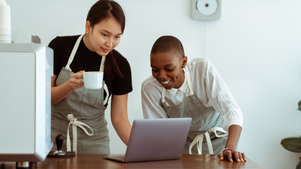 two people wearing aprons looking at a laptop and smiling 